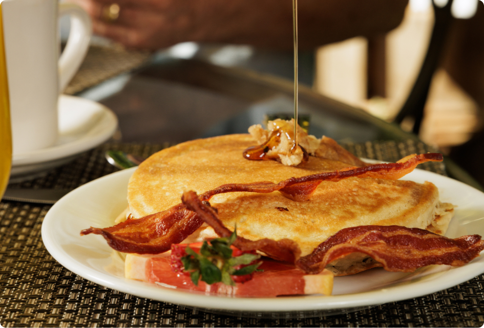 A plate with pancakes, bacon, and fruit is on a woven placemat. A person pours syrup, with a cup nearby.