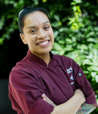 A person in a burgundy chef’s jacket smiles with folded arms against a lush green plant background, exuding confidence and warmth.