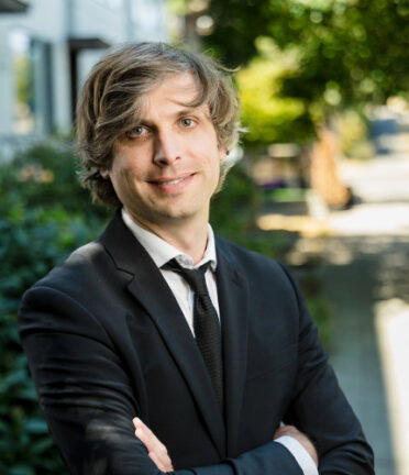 A person in a suit stands outdoors on a sunny day, with greenery and a sidewalk in the background.