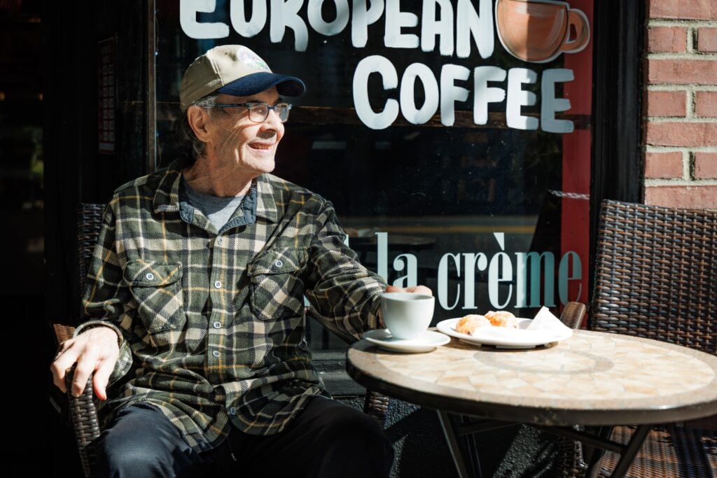 A person in a plaid shirt sits outside a cafe, enjoying coffee and pastries in the sunlight, next to a window advertising European coffee.